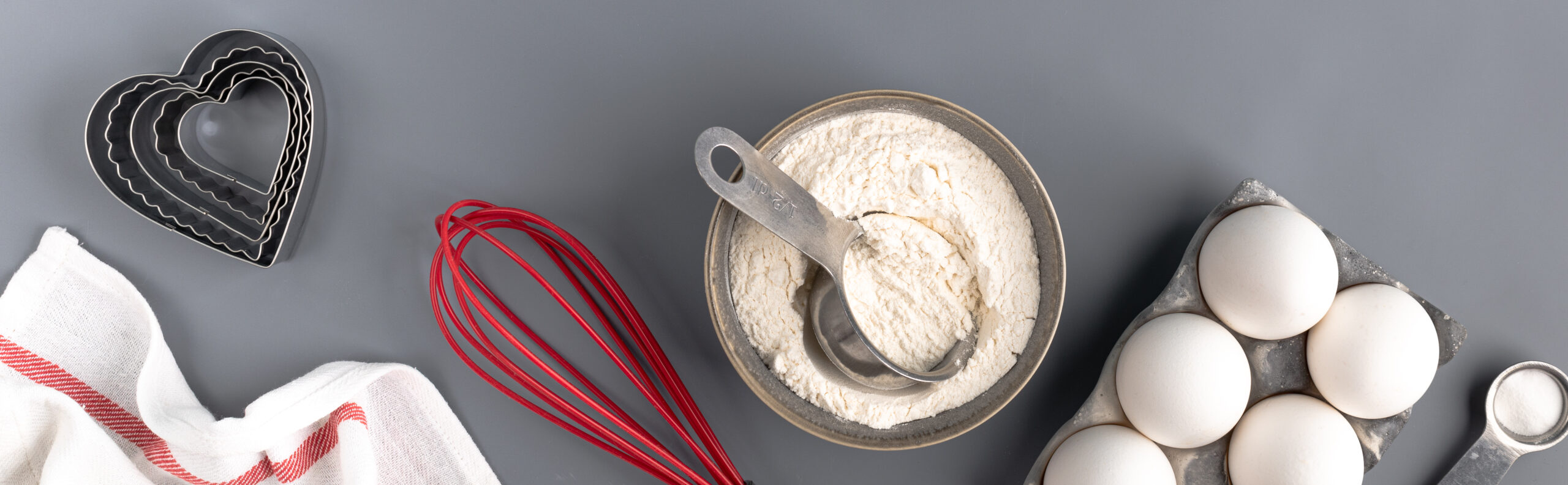 Ingredients for baking on a dark gray background: flour, eggs, baking powder, heart shape, whisk, textiles. top view. Copy space. Layout of a pie or cake recipe with cookies